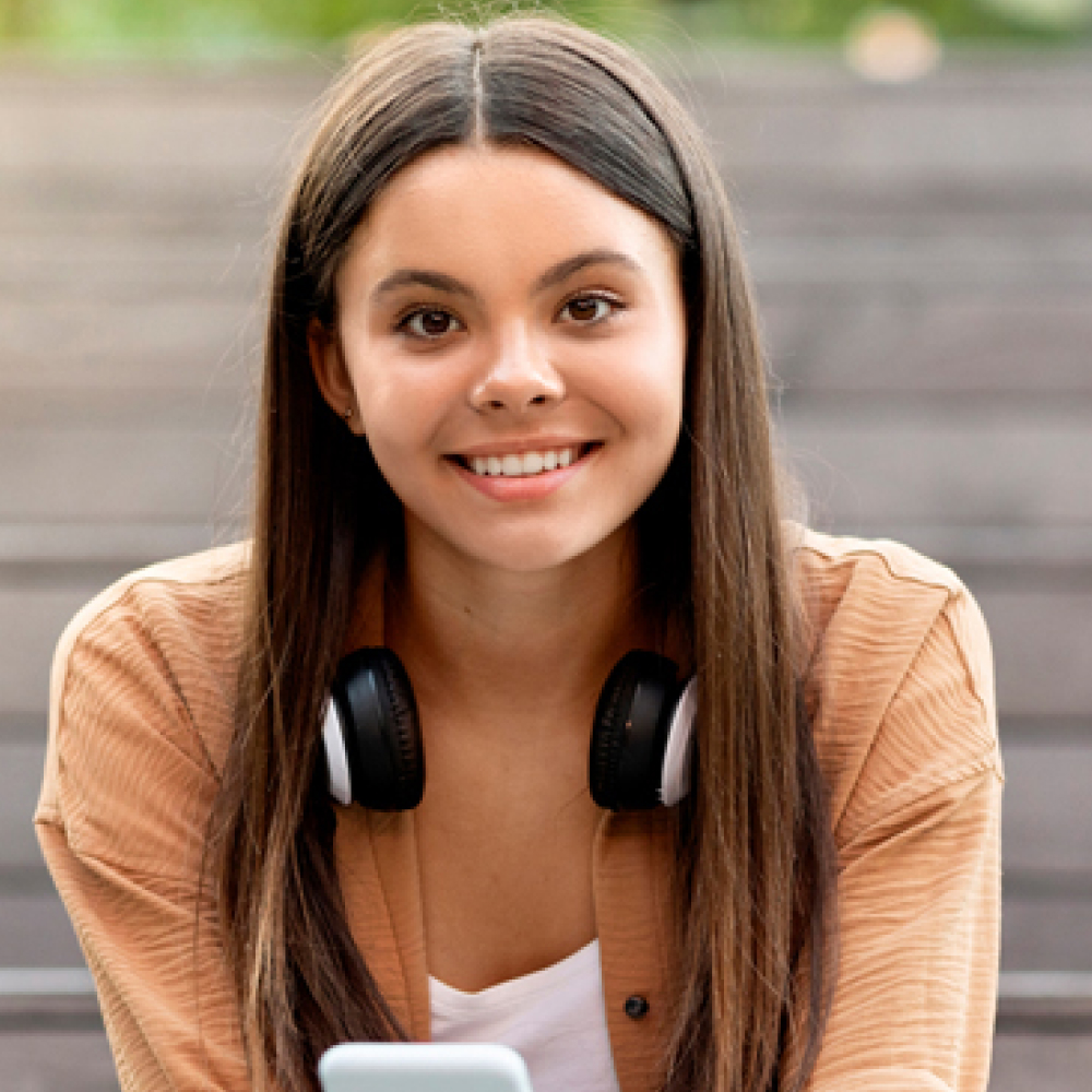 Smiling young woman with long brown hair wears black headphones and a tan jacket, posing outdoors against blurred steps with a friendly expression.
