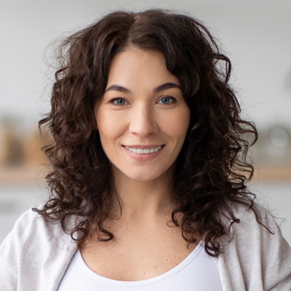 Smiling woman with long, curly dark hair and blue eyes, wearing a white top and cardigan, posed against a softly blurred background.