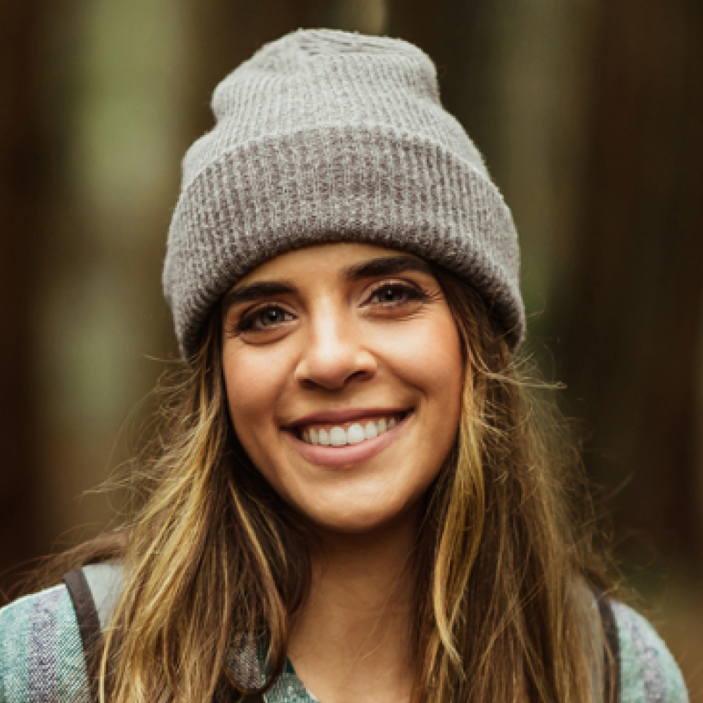 Smiling woman with long brown hair wears a gray knit beanie and outdoor jacket, warmly lit against a softly blurred forest background.