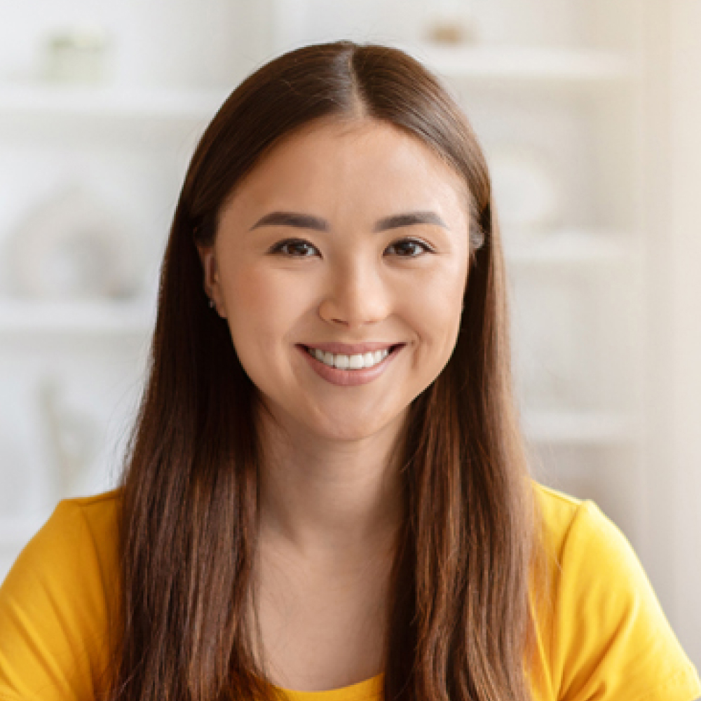 Smiling woman with long brown hair, wearing a bright yellow top, looks directly at the camera in a softly lit home, conveying warmth and confidence.